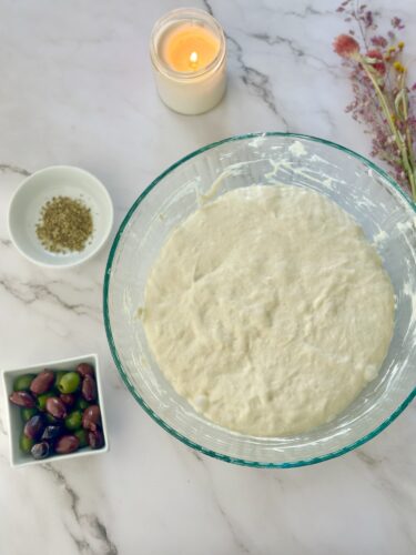 A glass bowl filled with dough resting on a marble countertop, surrounded by a small bowl of herbs, a dish of olives, a lit candle, and dried flowers.