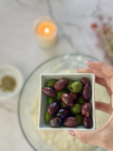 A hand holding a small white bowl filled with green and black olives, with a lit candle and ingredients blurred in the background.
