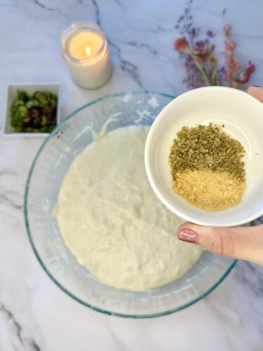 A hand holding a small bowl of spices above a large bowl of dough, with a lit candle and fresh herbs visible in the background.