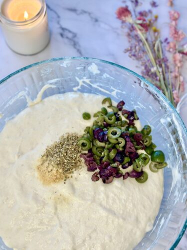 A clear glass bowl filled with a creamy mixture of dough, topped with chopped green olives, black olives, and a sprinkle of herbs, with a lit candle and dried flowers in the background.