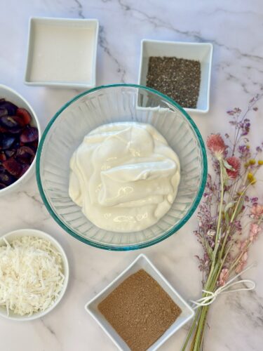 A glass bowl filled with creamy yogurt, surrounded by small bowls containing black chia seeds, shredded coconut, and brown sugar. There are also fresh plums and a bouquet of dried flowers on a marble countertop.