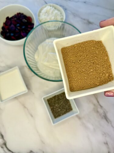 A close-up of a hand holding a dish of brown sugar, surrounded by various ingredients including a bowl of mixed fruits, a bowl of shredded coconut, and small dishes of cream and herbs on a marble countertop.