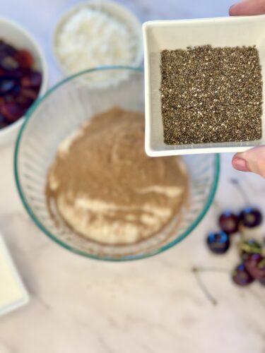 A hand holding a small dish of dark spice over a mixing bowl filled with dry ingredients, surrounded by bowls of fruit and a plate of butter.