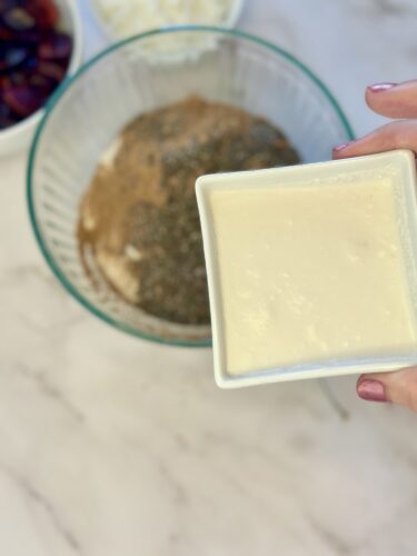 A hand holding a small square dish of creamy sauce above a glass bowl containing mixed dry ingredients.