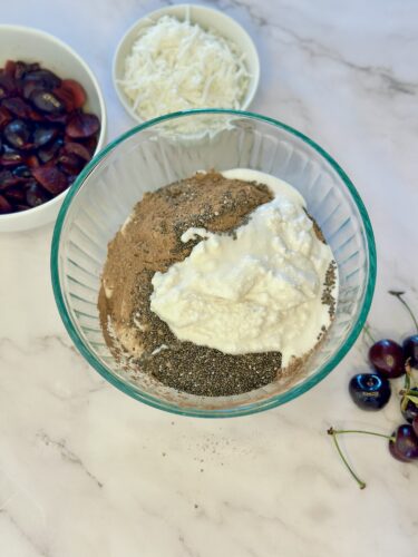 A glass bowl filled with a mixture of dry ingredients and yogurt, surrounded by bowls of shredded coconut and dried fruits, with fresh cherries on the side.