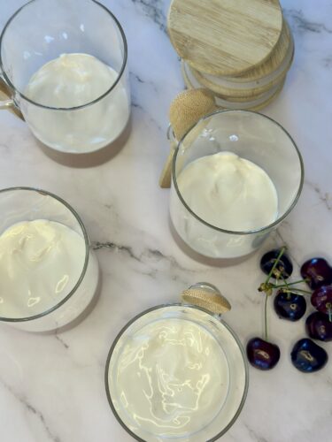Three glasses filled with a creamy dessert are placed on a marble surface, accompanied by a small wooden lid and a handful of cherries.