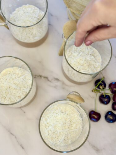 Four transparent cups filled with shredded coconut, with a hand sprinkling additional coconut over them. Fresh cherries are placed nearby on a marble surface.