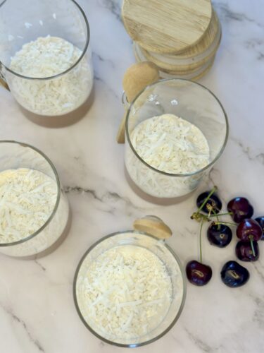 Four glass jars filled with shredded coconut, with a wooden lid and spoon nearby, and a bunch of cherries on the marble surface.