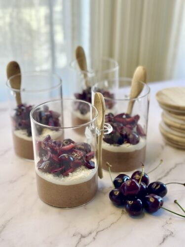 Four dessert glasses filled with chocolate mousse topped with cherries and whipped cream, accompanied by wooden spoons and a few fresh cherries on the side.