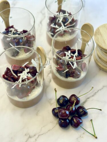 Four glass dessert cups filled with chocolate mousse, topped with cherries and shredded coconut. A cluster of cherries is placed on the marble surface beside the cups.