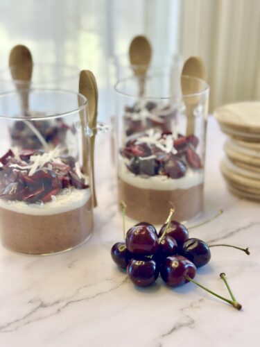 A close-up of dessert cups filled with layers of chocolate mousse, whipped cream, and topped with pieces of cherries and coconut flakes. A cluster of fresh cherries is placed in front of the cups.
