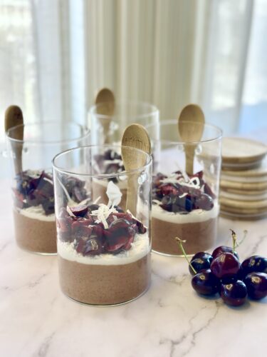 Four glass dessert cups layered with chocolate mousse and topped with cherries, coconut flakes, and wooden spoons, with fresh cherries in the foreground.