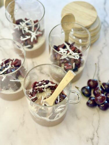 Four dessert cups with layers of cream, topped with cherries and shredded coconut, placed on a marble surface, with a wooden spoon in one cup and cherries beside the cups.