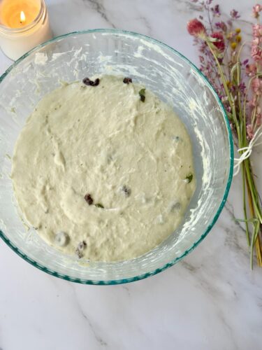 A glass mixing bowl filled with thick batter containing raisins and herbs, set on a marble countertop with a lit candle and dried flowers in the background.