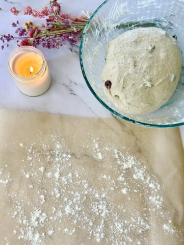 A glass bowl containing dough with herbs and a chocolate chip, placed next to a sheet of parchment paper sprinkled with flour and a lit candle, accompanied by dried flowers.