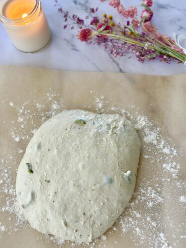 A flattened ball of dough on a sheet of parchment paper, surrounded by a sprinkle of flour. A lit candle and dried flowers are visible in the background.