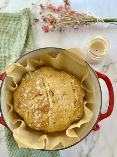 Freshly baked round bread in a red pot lined with parchment paper, surrounded by a green kitchen cloth and a candle, with dried flowers in the background.
