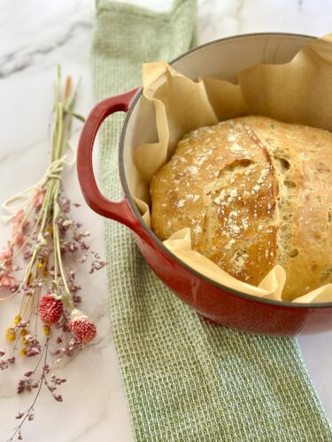 A freshly baked loaf of bread in a red Dutch oven, lined with parchment paper, placed on a green kitchen towel next to a bouquet of dried flowers.