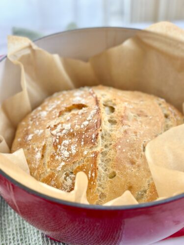 A freshly baked round loaf of bread resting in a red pot lined with parchment paper.