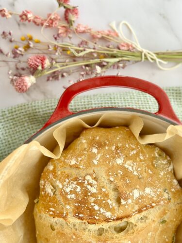 A freshly baked loaf of bread in a red Dutch oven, with a decorative touch of dried flowers in the background.
