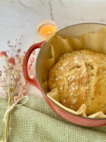 A freshly baked loaf of bread in a red cast-iron pot lined with parchment paper, surrounded by dried flowers and a lit candle on a marble surface.