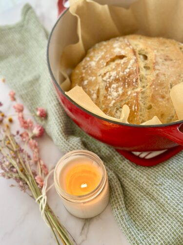 Freshly baked bread in a red Dutch oven with parchment paper, surrounded by dried flowers and a lit candle on a marble surface.