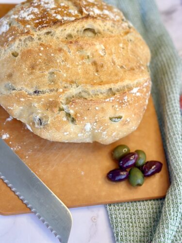 Freshly baked rustic olive bread on a wooden cutting board with a serrated knife and a few olives beside it.