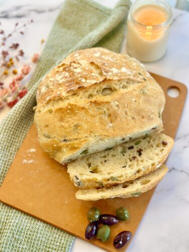 A freshly baked loaf of artisan bread on a wooden cutting board, with slices cut and a few olives beside it. A soft green cloth and a lit candle are in the background.