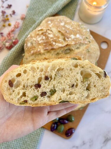A hand holding a sliced piece of artisan bread showing a crumb with green and purple olives, set against a background of a whole loaf and decorative elements.