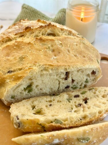 A loaf of homemade bread with green olives and sundried tomatoes, partially sliced on a wooden cutting board, beside a lit candle.