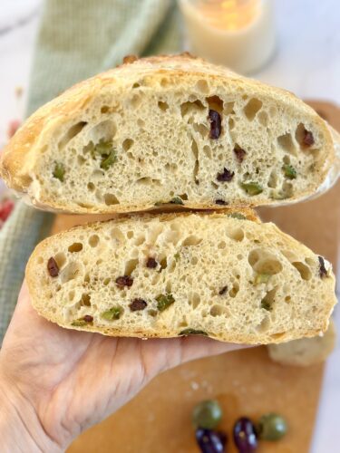 Close-up of a hand holding a halved loaf of bread featuring a soft, airy crumb with pieces of green and black olives embedded throughout.