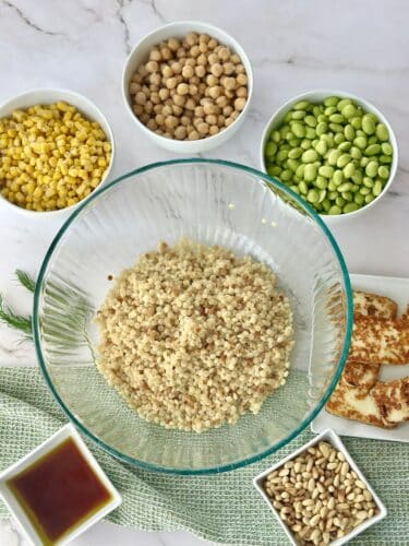 A bowl of cooked couscous surrounded by various ingredients including corn, chickpeas, edamame, pine nuts, and pieces of haloumi cheese, with a small bowl of dressing on the side.