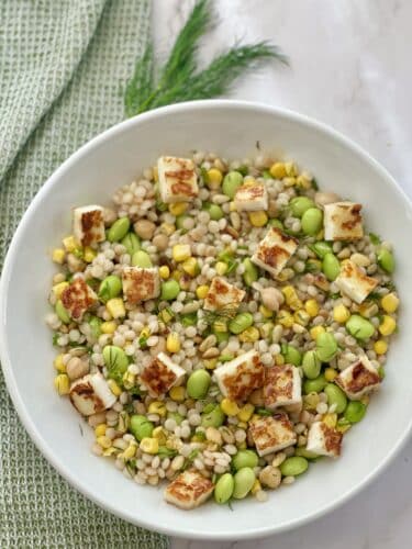 A bowl of grain salad featuring pearl couscous, green peas, corn, and cubed grilled halloumi cheese, garnished with fresh herbs.