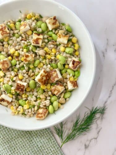 A bowl of barley salad with grilled halloumi cheese, green edamame, sweet corn, and fresh dill, served on a marble surface.