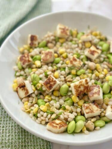 A bowl of barley salad topped with sautéed halloumi cheese, mixed with green peas, corn, and chickpeas.