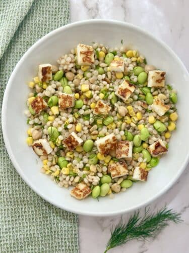 A colorful salad featuring barley, corn, and edamame, topped with cubes of lightly browned halloumi cheese and fresh dill, served in a white bowl.