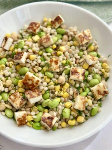 A bowl of mixed grain salad featuring cooked pearls, green edamame, corn, and cubes of grilled cheese.
