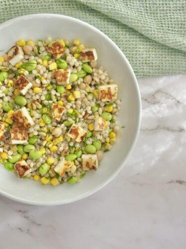 A bowl of mixed grains and vegetables including edamame, corn, and chickpeas topped with grilled cheese cubes, served on a marble surface with a textured green cloth in the background.