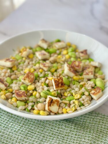 A bowl of salad featuring pearl couscous, green edamame, corn, and grilled halloumi cheese, served on a textured green placemat.