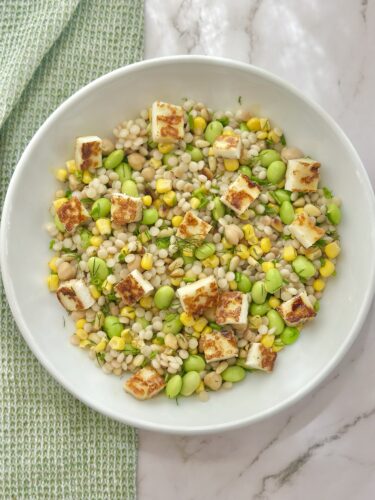 A bowl of mixed grain salad featuring pearl barley, corn, edamame, and white beans, topped with grilled cheese cubes and garnished with fresh herbs.