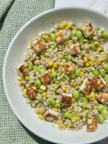 A bowl of pearl couscous salad topped with grilled halloumi cheese, edamame beans, sweet corn, and herbs.