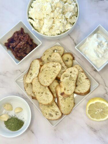 A platter of sliced toasted bread surrounded by small bowls containing sun-dried tomatoes, cream cheese, crumbled feta cheese, garlic cloves, lemon, and dried herbs.