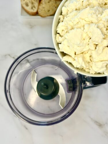 A food processor with a clear bowl and a black base, positioned alongside a bowl filled with ricotta cheese and a plate of sliced bread in the background.