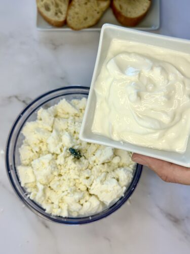 A hand holding a bowl of sour cream above a dish of crumbled cottage cheese, with slices of bread in the background.