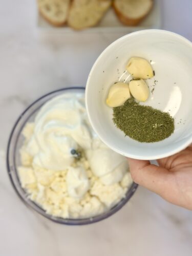 A hand holding a bowl with garlic cloves and dried herbs above a food processor filled with yogurt and cheese.
