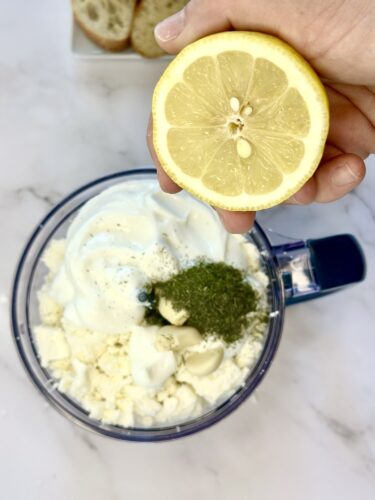 A hand holding a half lemon above a food processor filled with yogurt, herbs, and cottage cheese, with slices of bread in the background.