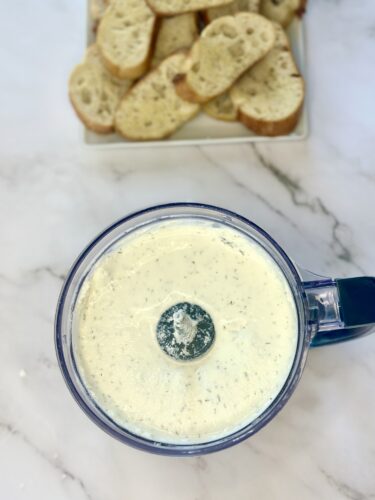 Top view of a food processor filled with a creamy sauce, with slices of bread in the background.