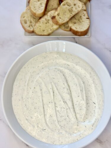 A creamy dip in a white bowl with a smooth texture, accompanied by slices of bread on a plate in the background.