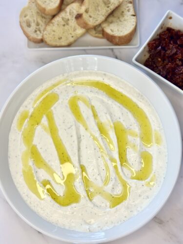 A bowl of creamy yogurt dip drizzled with olive oil, accompanied by slices of bread and a small dish of red sauce.