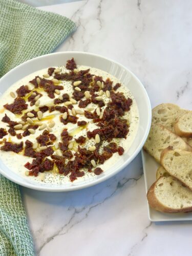 A white bowl filled with creamy dip topped with sun-dried tomatoes, pine nuts, and herbs, accompanied by slices of crusty bread on a plate.